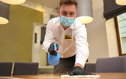 custodian staff cleaning surface of table with face mask, gloves and googles on.
